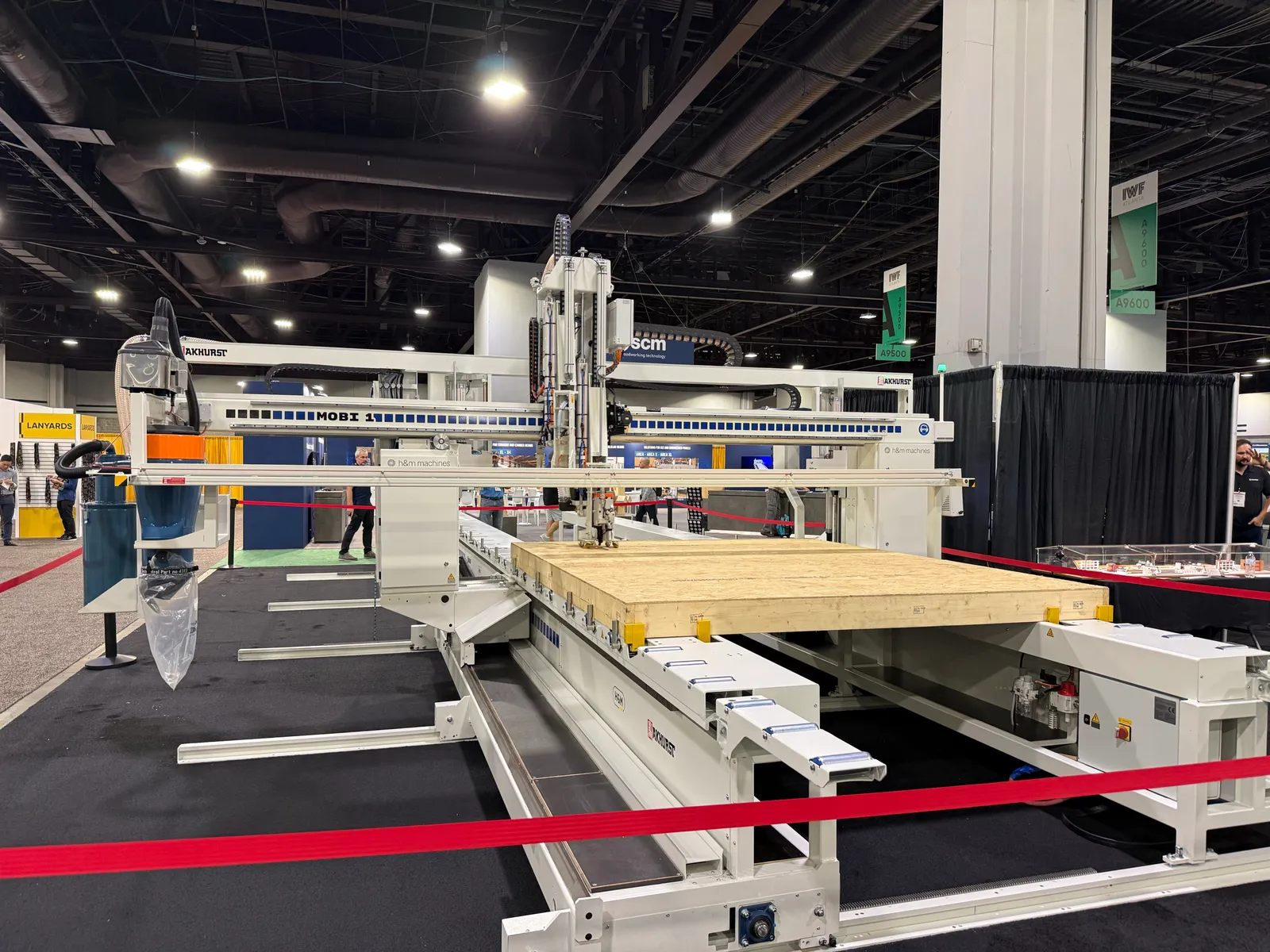 A large Akhurst MOBI 1 six-axis CNC gantry machine on the show floor, its head positioned over a wood floor cassette on the infeed table, with the SCM booth in the background.