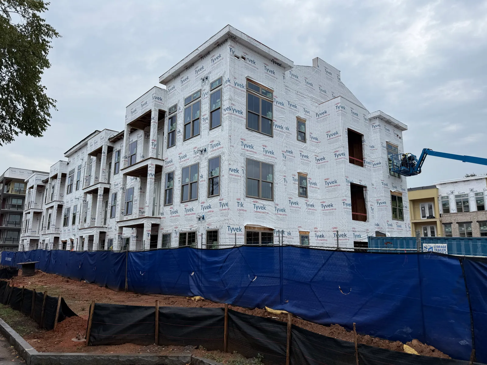A multi-story wood-framed multifamily building under construction, wrapped in DuPont Tyvek CommercialWrap, behind a blue site fence, with a worker on a boom lift.