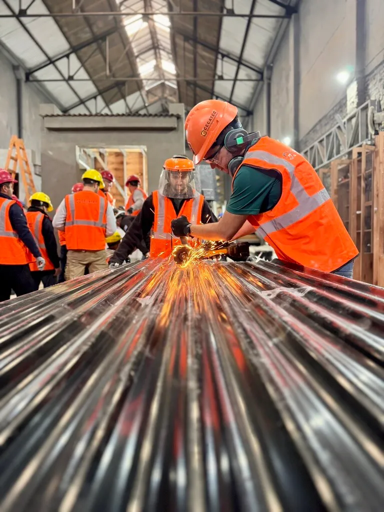 A course participant in a face shield uses an angle grinder to cut a steel framing profile, sparks flying, on the CECATEC workshop floor.