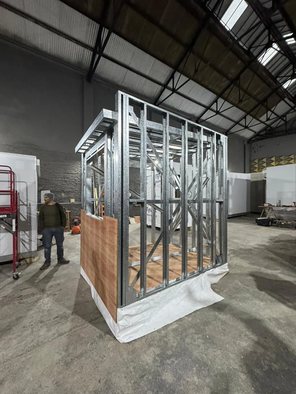 A completed light gauge steel wall frame with a plywood floor and partial sheathing, assembled by the course, standing on the workshop floor.